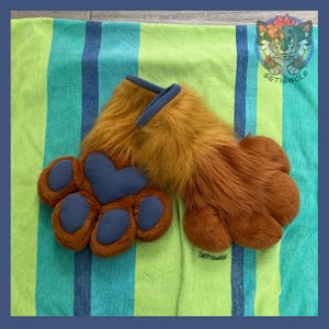 May include: A pair of brown faux fur paw gloves featuring dark blue fleece paw pads and a heart-shaped palm pad. The cuffs are trimmed with dark blue fabric. A small logo with a stylized wolf character and the text Setiswolf is visible.