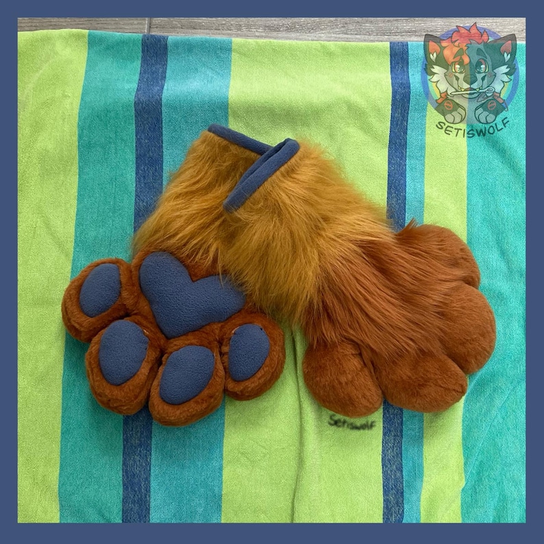 May include: A pair of brown faux fur paw gloves featuring dark blue fleece paw pads and a heart-shaped palm pad. The cuffs are trimmed with dark blue fabric. A small logo with a stylized wolf character and the text Setiswolf is visible.