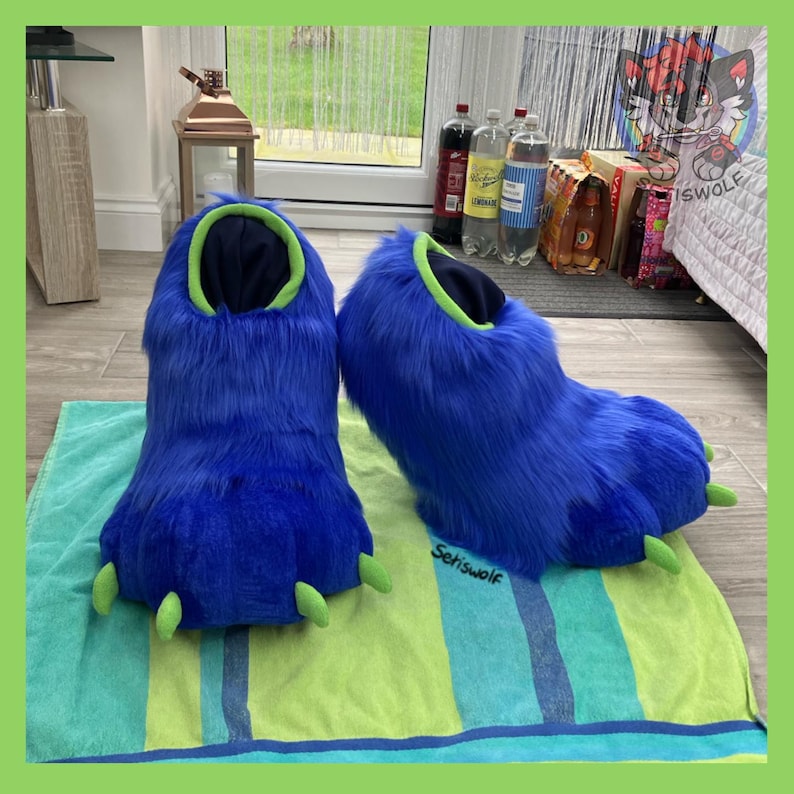 May include: A pair of blue furry monster feet slippers with green claws and trim. The slippers are resting on a green and blue striped towel. The text "Setiswolf" is visible on the towel.