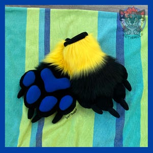 May include: A pair of plush animal paw gloves featuring long black and bright yellow faux fur. The palm side is made of black fabric with four large, rounded blue paw pads and black claws extending from the tips of each finger section.
