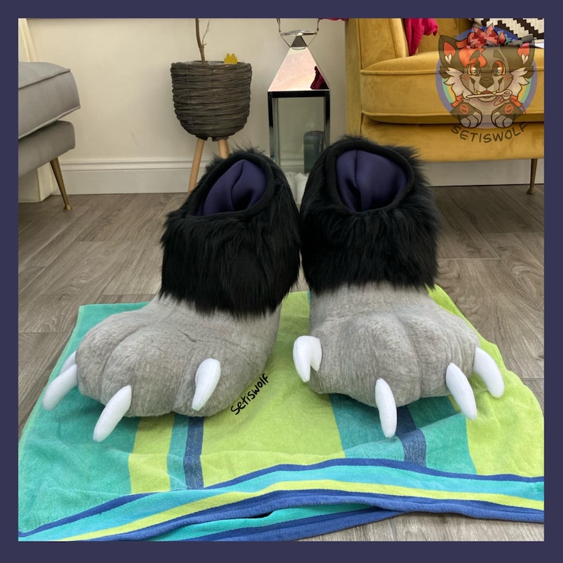 May include: A pair of novelty slippers shaped like large gray paws with white claws. The top of the slippers are black and fluffy, with a dark blue interior. The slippers are displayed on a green and blue striped towel.
