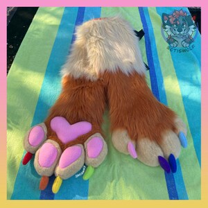 May include: A pair of faux fur hand paws featuring long orange and cream-colored fur. The paws have large pink heart-shaped palm pads, smaller pink finger pads, and multi-colored claws in red, orange, yellow, lime green, purple, and blue.