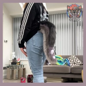 May include: A gray and purple faux fur tail with white accents is attached to the back of a person wearing blue jeans. The tail is long and fluffy, with a gradient of colors. The tail is a costume accessory.