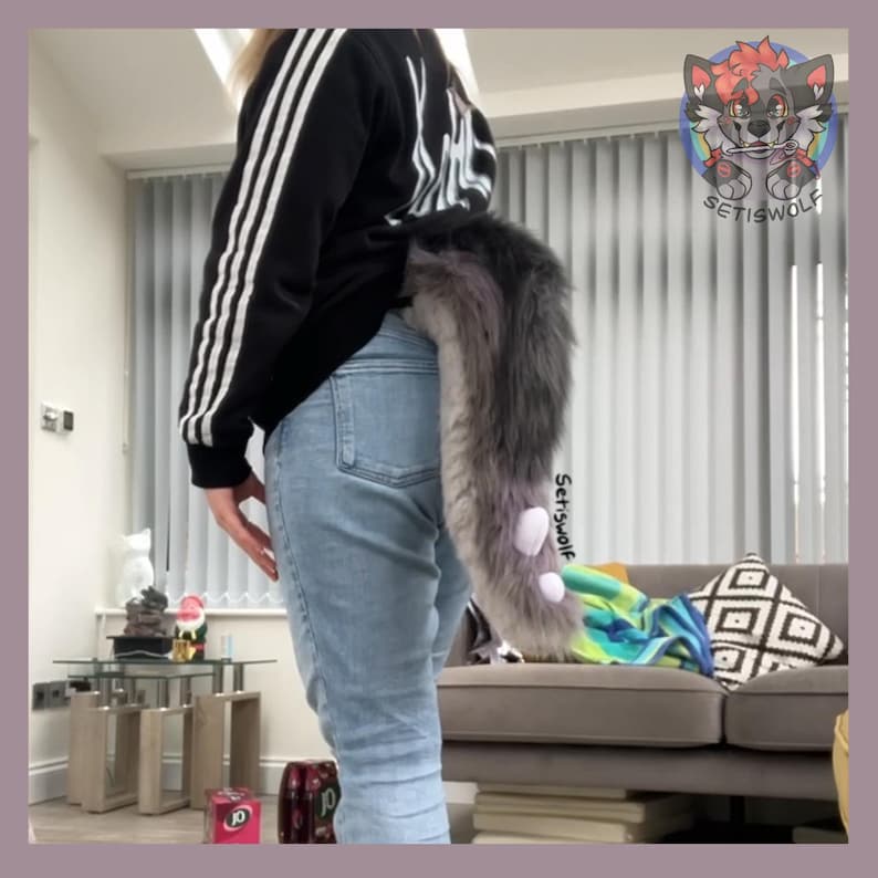 May include: A gray and purple faux fur tail with white accents is attached to the back of a person wearing blue jeans. The tail is long and fluffy, with a gradient of colors. The tail is a costume accessory.