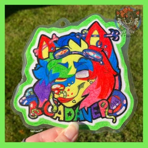 K&ouml;nnte beinhalten: Ein transparenter Acryl-Anh&auml;nger mit einer farbenfrohen, handgezeichneten Illustration einer stilisierten anthropomorphen Kreatur mit rotem, blauem und gelbem Fell, die eine Schutzbrille tr&auml;gt. Der Text CADAVER-D steht in bunten Buchstaben.