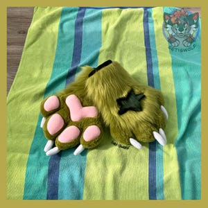 May include: A pair of olive green faux fur paw gloves featuring soft pink padded paw pads, white fabric claws, and a dark green star-shaped patch on the back of the hand. The gloves are textured with long synthetic fibers and have a plush appearance.