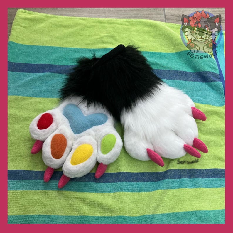 May include: A pair of plush animal paw gloves featuring black faux fur at the wrists and white faux fur on the palms. The palms display a light blue heart-shaped pad, four colorful oval pads in red, orange, yellow, and green, and pink claws.