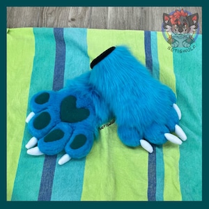 May include: A pair of bright blue faux fur paw gloves featuring dark teal paw pads and a heart-shaped center pad. The gloves have white claws and a black cuff opening. A small logo with the text Setiswolf is visible on the fur surface.