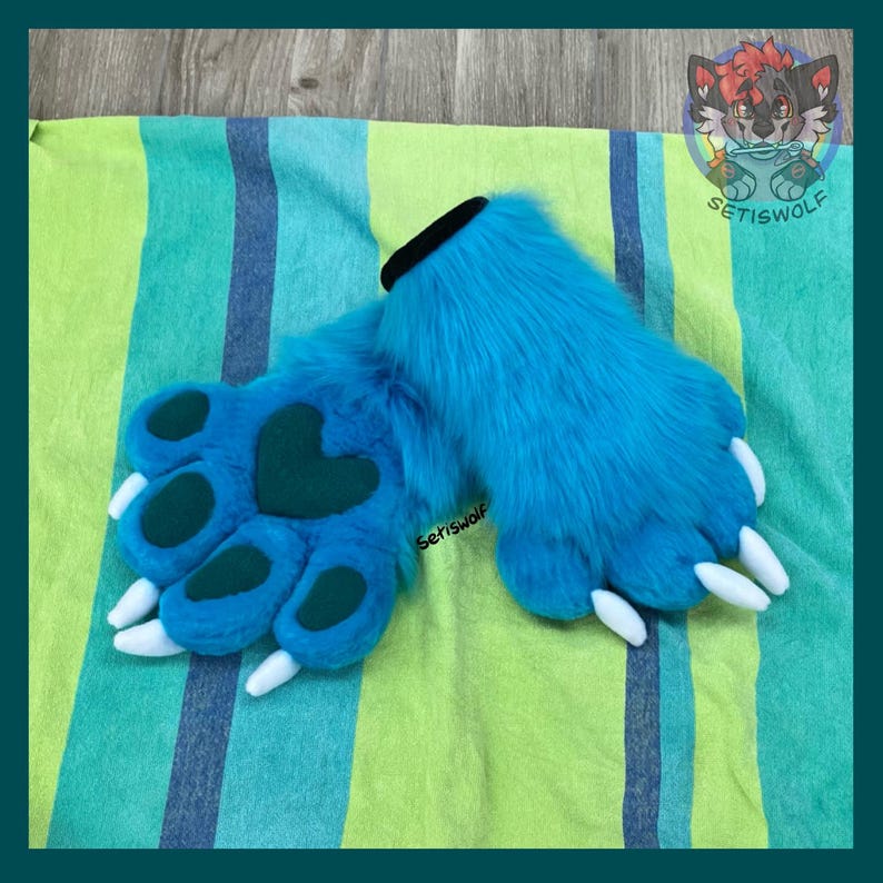 May include: A pair of bright blue faux fur paw gloves featuring dark teal paw pads and a heart-shaped center pad. The gloves have white claws and a black cuff opening. A small logo with the text Setiswolf is visible on the fur surface.