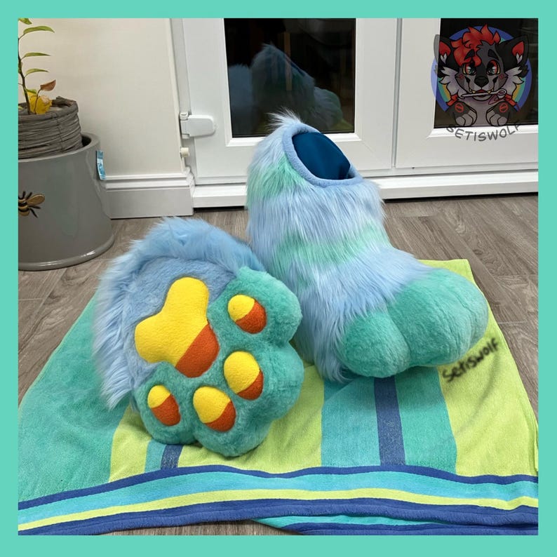 May include: A pair of furry blue and green paw-shaped costume feet with yellow and orange paw pads. The paws are resting on a striped green and blue towel. The costume feet are designed to resemble a furry animal.