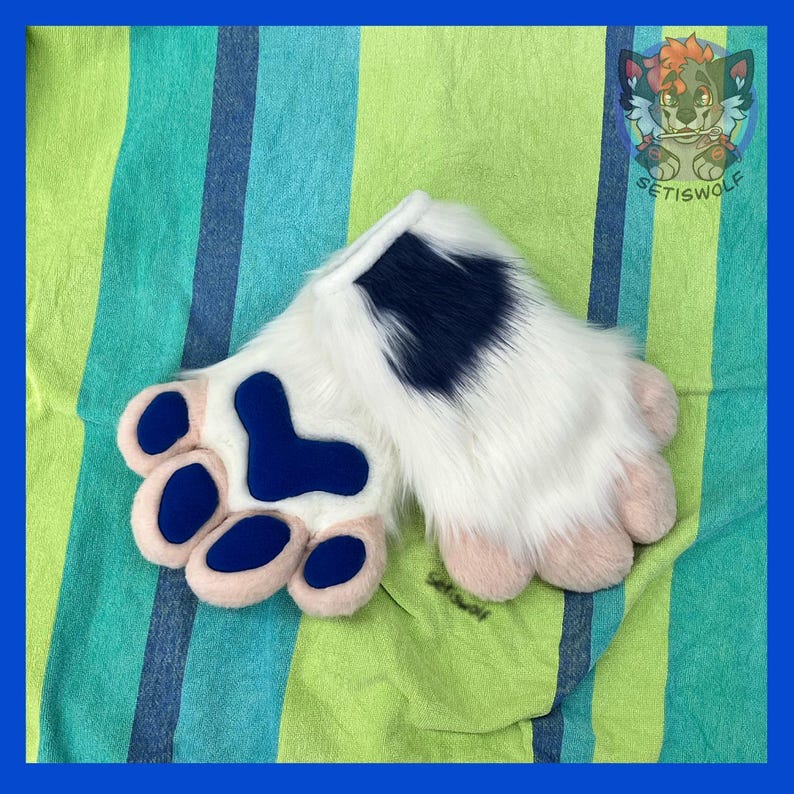 May include: A pair of white faux fur hand paws featuring dark blue paw pads and a dark blue patch on the wrist area. The fingers are tipped with light pink fabric, and the design includes a logo in the corner with the text SETISWOLF.