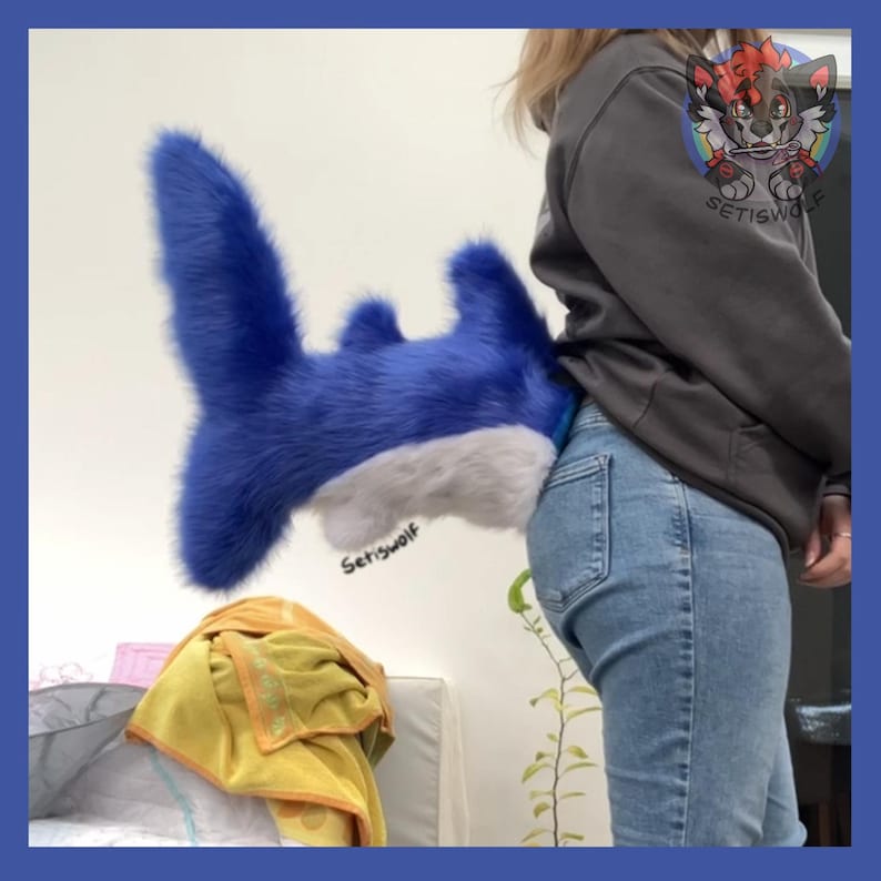 May include: A plush shark tail in shades of blue and white. The tail is attached to a person's jeans. The shark tail is made of soft, fluffy material. The text "Setiswolf" is visible on the tail.