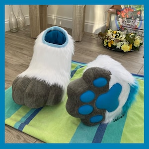 May include: A pair of furry, white and gray paw-shaped costume feet. The feet have a blue interior and paw pads. The bottoms are gray with blue paw pads. The feet are on a striped green and blue towel.