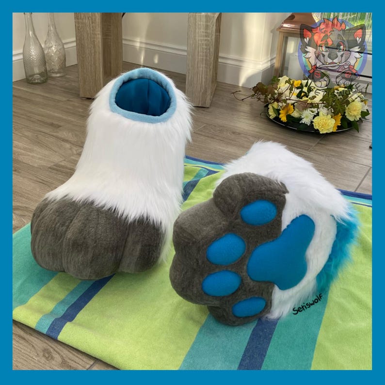 May include: A pair of furry, white and gray paw-shaped costume feet. The feet have a blue interior and paw pads. The bottoms are gray with blue paw pads. The feet are on a striped green and blue towel.