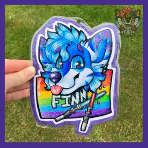 K&ouml;nnte beinhalten: Ein transparenter Acryl-Anh&auml;nger mit einem blau-wei&szlig;en anthropomorphen Wolfscharakter mit rosa Zunge. Die Figur ist &uuml;ber dem Namen FINN in wei&szlig;en Buchstaben platziert, vor einem Regenbogenhintergrund mit zwei gekreuzten Lichtschwertern darunter.