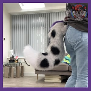 May include: A white and black spotted faux fur tail with a purple tip. The tail is attached to a person wearing blue jeans. The tail is a furry accessory.