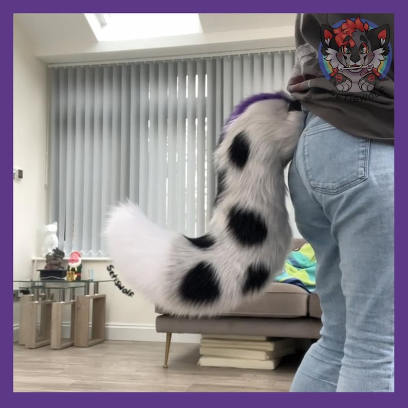 May include: A white and black spotted faux fur tail with a purple tip. The tail is attached to a person wearing blue jeans. The tail is a furry accessory.
