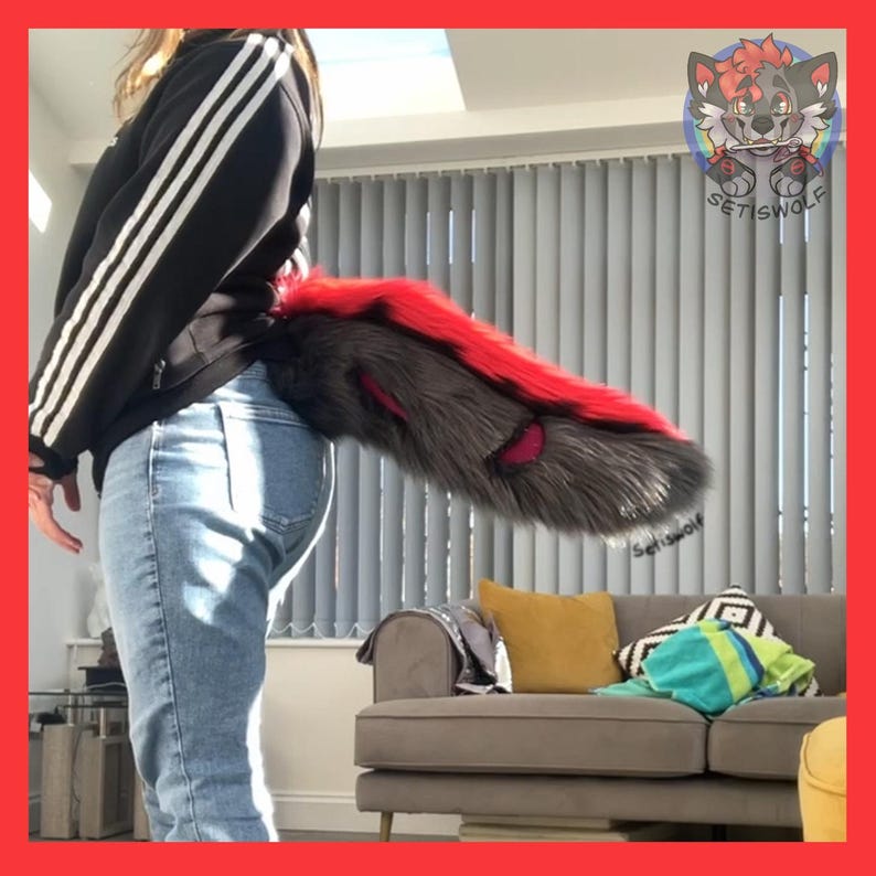 May include: A furry tail in shades of gray, red, and pink, attached to the back of a person wearing blue jeans. The tail is long and fluffy, with a red stripe along the top. The tail is a costume accessory.