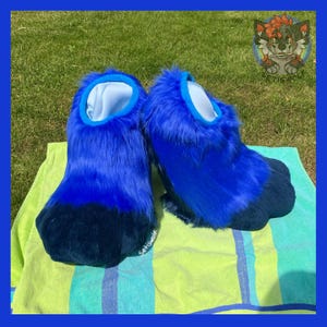 May include: A pair of blue furry paw-shaped costume feet. The feet are a gradient of blue, with a dark blue paw pad and a light blue interior. The feet are resting on a striped towel.