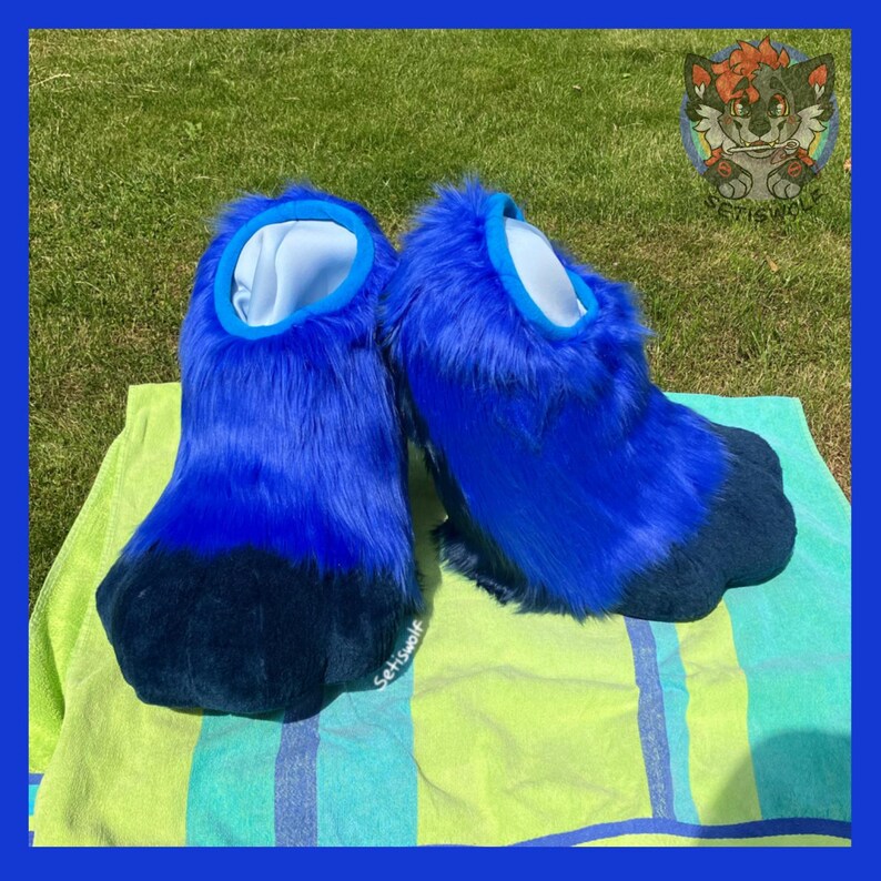 May include: A pair of blue furry paw-shaped costume feet. The feet are a gradient of blue, with a dark blue paw pad and a light blue interior. The feet are resting on a striped towel.