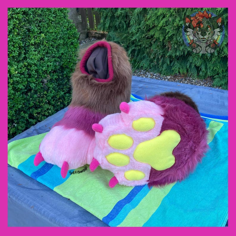 May include: A pair of furry, plush paws and a leg, in shades of brown, pink, and purple. The paw pads are yellow. The items are on a striped towel with a green and blue pattern. The leg has a pink trim.