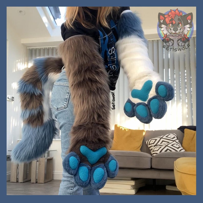 May include: A pair of long, plush faux fur arm sleeves and a matching tail. One sleeve is brown with a blue paw pad, the other is white with a blue paw pad. The tail features alternating bands of brown, white, and blue fur. A logo is visible.