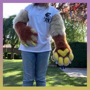 May include: A pair of long, plush faux fur arm sleeves featuring a gradient of tan and brown colors. The attached paw-shaped gloves have yellow heart-shaped pads, yellow toe pads, and multi-colored claws in red, orange, yellow, blue, and pink.