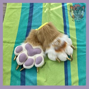 May include: A pair of plush animal paw gloves featuring long tan and white faux fur, soft lavender fabric paw pads, and pointed gold-colored claws. The wrist openings are trimmed with a yellow band, and the design includes a cartoon wolf logo.