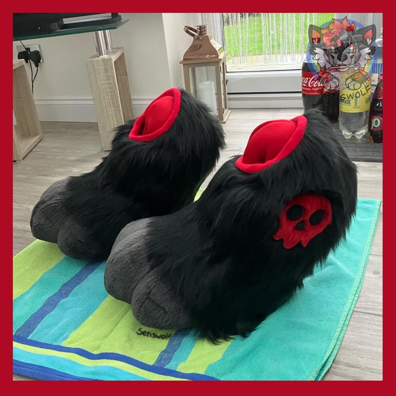 May include: A pair of black furry slippers with red interiors and gray paw pads. Each slipper features a red skull applique on the side. The slippers are displayed on a striped green and blue towel.