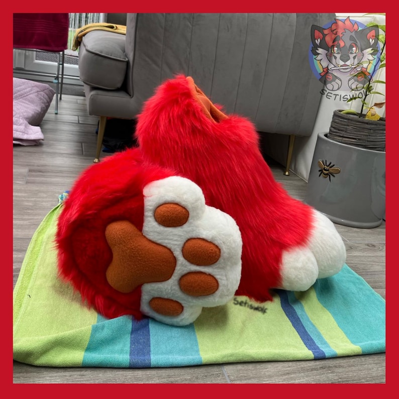 May include: A pair of red furry paws and feet, with white and orange paw pads. The feet are long and fluffy, with white tips. The paws are resting on a striped green and blue towel. The image is framed with a red border.