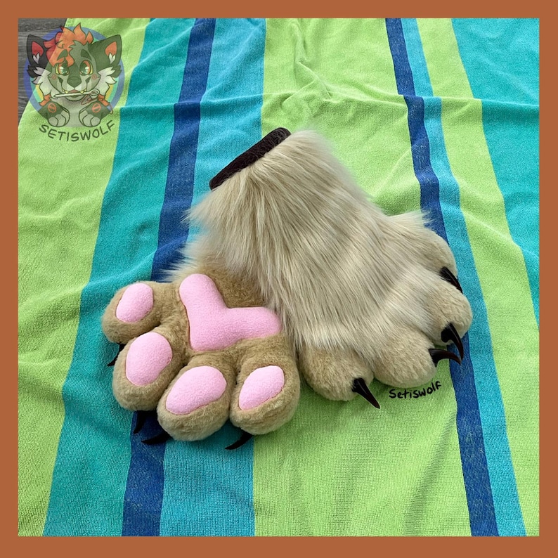 May include: A pair of beige faux fur hand paws featuring soft pink fabric paw pads and sharp black claws. The wrist opening is lined with dark brown fabric. The design includes a small circular logo in the corner with the text SETISWOLF.
