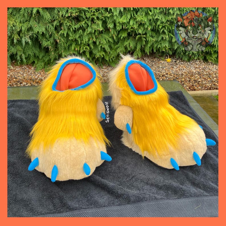 May include: A pair of furry yellow and tan paw-shaped slippers. The slippers have blue claws, orange interiors, and blue trim around the opening. The slippers are on a dark gray surface.