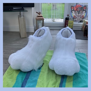 May include: A pair of white furry paw-shaped slippers. The slippers have a rounded toe and a fluffy texture, resembling animal paws. They are designed to be worn indoors and are placed on a green and blue striped towel.