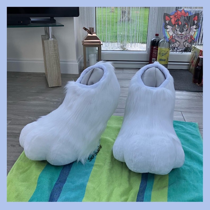 May include: A pair of white furry paw-shaped slippers. The slippers have a rounded toe and a fluffy texture, resembling animal paws. They are designed to be worn indoors and are placed on a green and blue striped towel.