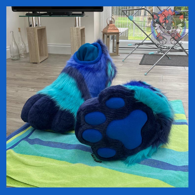 May include: A pair of blue and teal furry paw-shaped feet, likely for a costume. The paws have a dark blue base with lighter blue and teal accents. The soles feature large, dark blue paw pads. The feet are resting on a striped towel.
