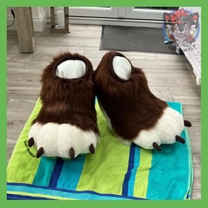 May include: A pair of brown and white furry paw slippers with black claws. The slippers have a white interior and are resting on a green and blue striped towel. The slippers are designed to resemble animal paws.