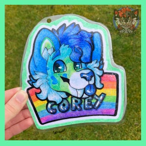 K&ouml;nnte beinhalten: Ein durchsichtiger Acryl-Anh&auml;nger mit einer farbenfrohen Illustration eines blau-gr&uuml;nen anthropomorphen Wolfscharakters mit gro&szlig;en Augen und herausgestreckter Zunge. Der Name COREY steht in schwarzen Buchstaben auf einem Regenbogenstreifen.