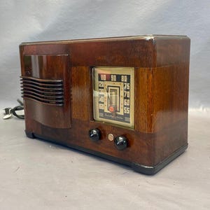 Może przedstawiać: Antyczne drewniane radio z brązową drewnianą obudową i srebrną płytą czołową. Radio ma tarczę z liczbami od 55 do 110. Radio ma dwa pokrętła z przodu.