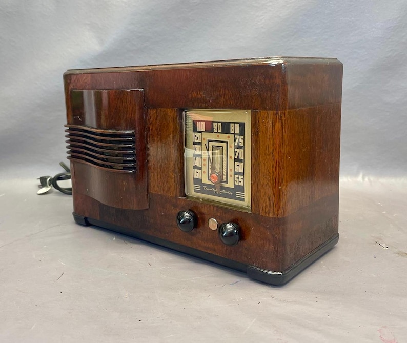 Może przedstawiać: Antyczne drewniane radio z brązową drewnianą obudową i srebrną płytą czołową. Radio ma tarczę z liczbami od 55 do 110. Radio ma dwa pokrętła z przodu.