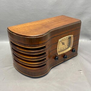 May include: A vintage wooden radio with a gold-colored dial and black knobs. The radio has a wood grain finish and a speaker grille with five horizontal slats.