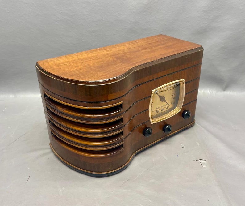 May include: A vintage wooden radio with a gold-colored dial and black knobs. The radio has a wood grain finish and a speaker grille with five horizontal slats.