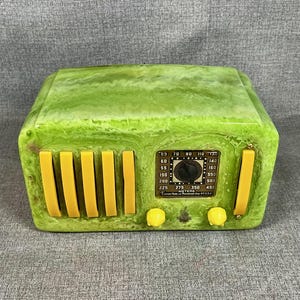 May include: A retro-style radio in a marbled green finish with yellow detailing. The radio has a dial, speaker bars, and control knobs. The face of the radio has a label with numbers and text.