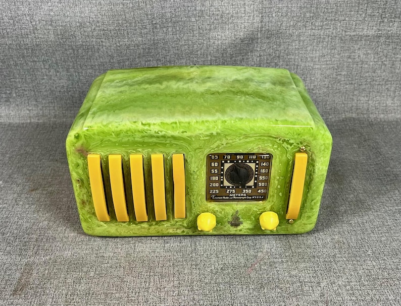 May include: A retro-style radio in a marbled green finish with yellow detailing. The radio has a dial, speaker bars, and control knobs. The face of the radio has a label with numbers and text.