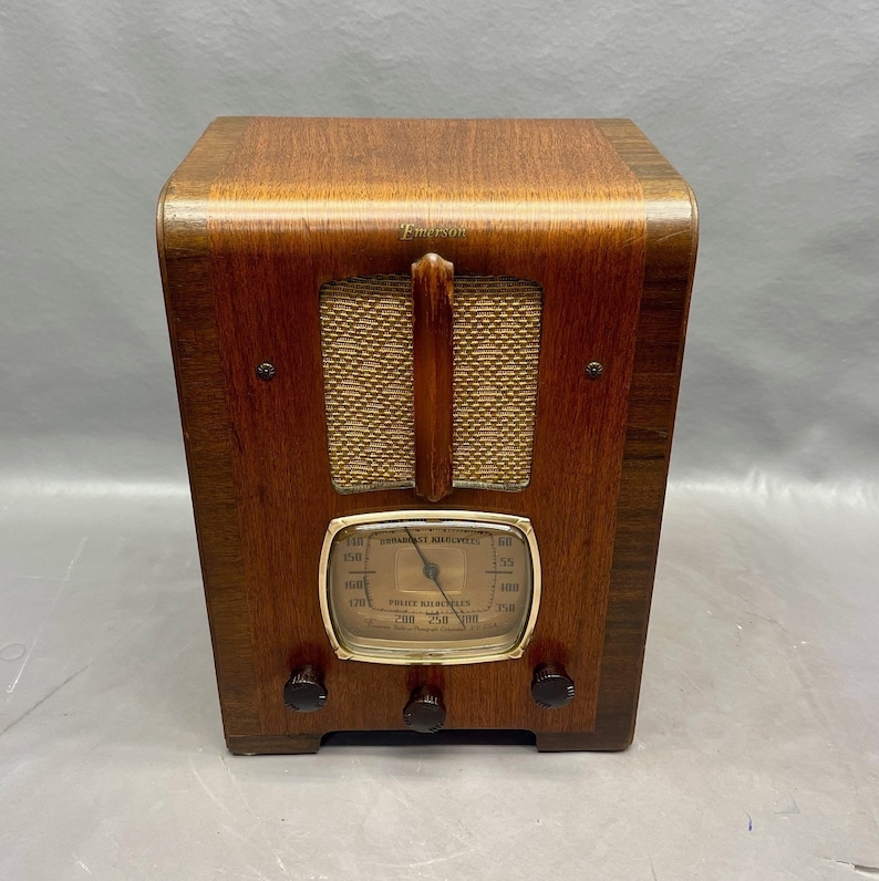 May include: A vintage wooden Emerson radio with a gold-colored dial and a woven speaker grille. The radio has a brown wooden cabinet with a black knob on each side.