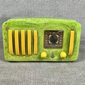 May include: A green and yellow decorative radio with a vintage design. The radio features yellow vertical bars, knobs, and a dial with numbers and the word "METERS". The radio has the text "FILOCYCLE" on the dial.