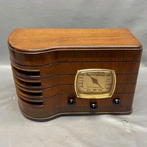 May include: A vintage wooden radio with a round dial and a gold-colored face. The dial has the text "Broadcast Wavelengths" and "Police Frequencies" printed on it. The radio has a dark wood finish and a curved design.