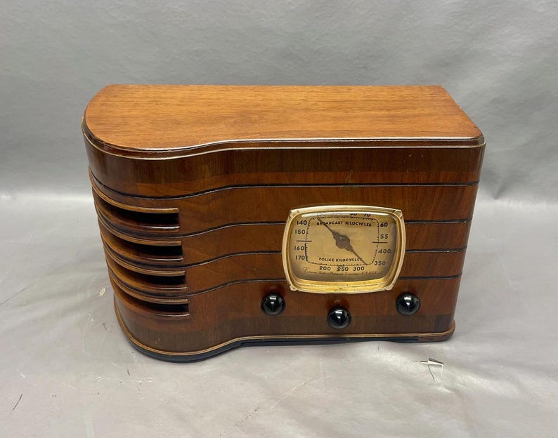 May include: A vintage wooden radio with a round dial and a gold-colored face. The dial has the text "Broadcast Wavelengths" and "Police Frequencies" printed on it. The radio has a dark wood finish and a curved design.