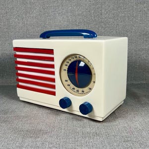 Pode incluir: Um r&aacute;dio de estilo vintage com corpo creme, listras vermelhas e al&ccedil;a azul. Possui um mostrador circular e dois bot&otilde;es azuis. O design lembra a est&eacute;tica de meados do s&eacute;culo. Dimens&otilde;es: aproximadamente 20 cm de largura.