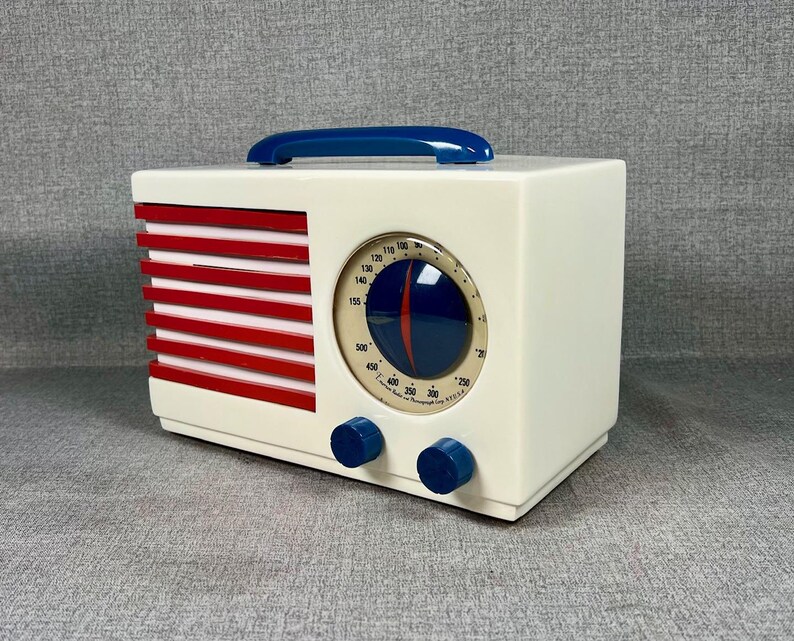 Pode incluir: Um r&aacute;dio de estilo vintage com corpo creme, listras vermelhas e al&ccedil;a azul. Possui um mostrador circular e dois bot&otilde;es azuis. O design lembra a est&eacute;tica de meados do s&eacute;culo. Dimens&otilde;es: aproximadamente 20 cm de largura.