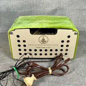May include: A vintage Emerson Radio & Phono Corp. radio with a light green top and a beige front panel. The front panel has a series of circular holes and the company's logo and text. Black and brown cords with a plug are in the foreground.
